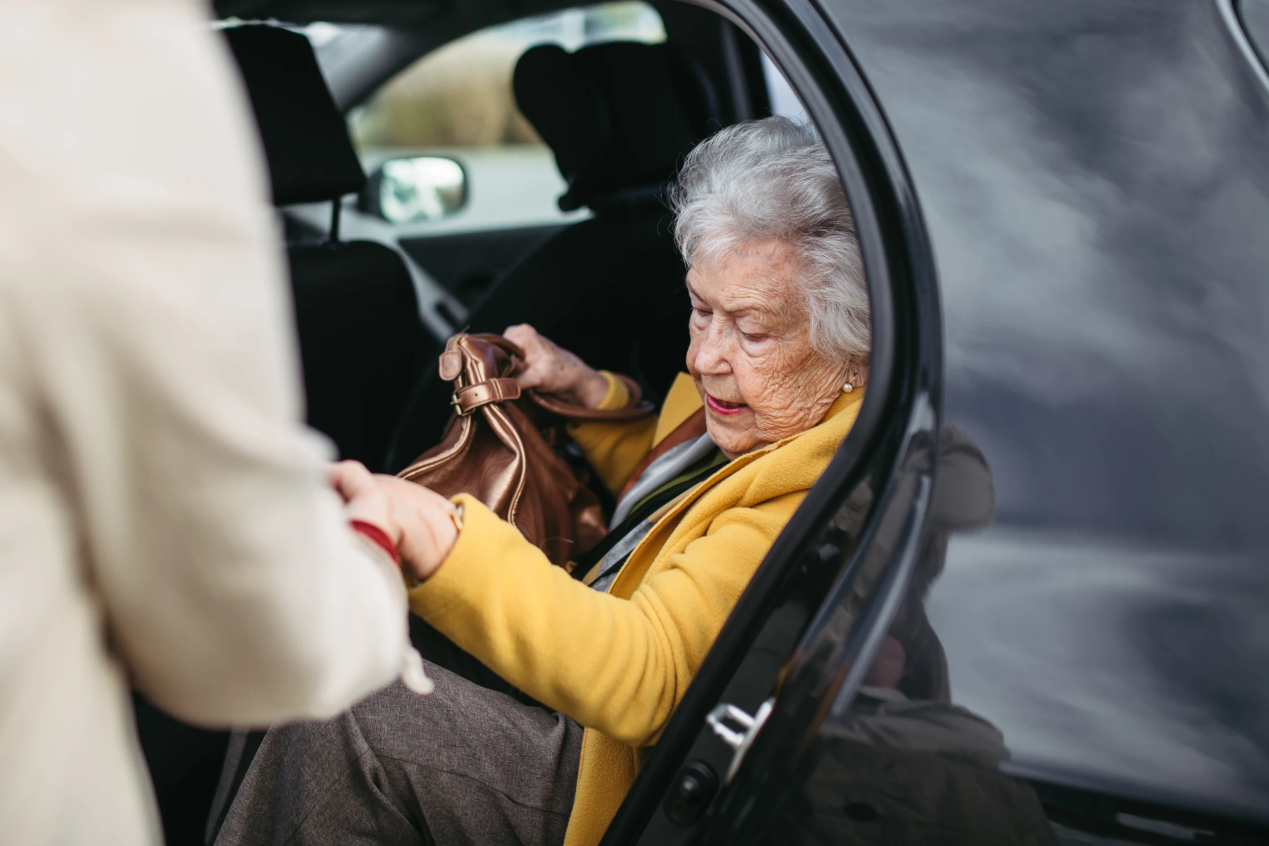 Senior lady getting out of the car, caregiver helping her, holding her hands. Elderly woman has problem with standing up from the car back seat.